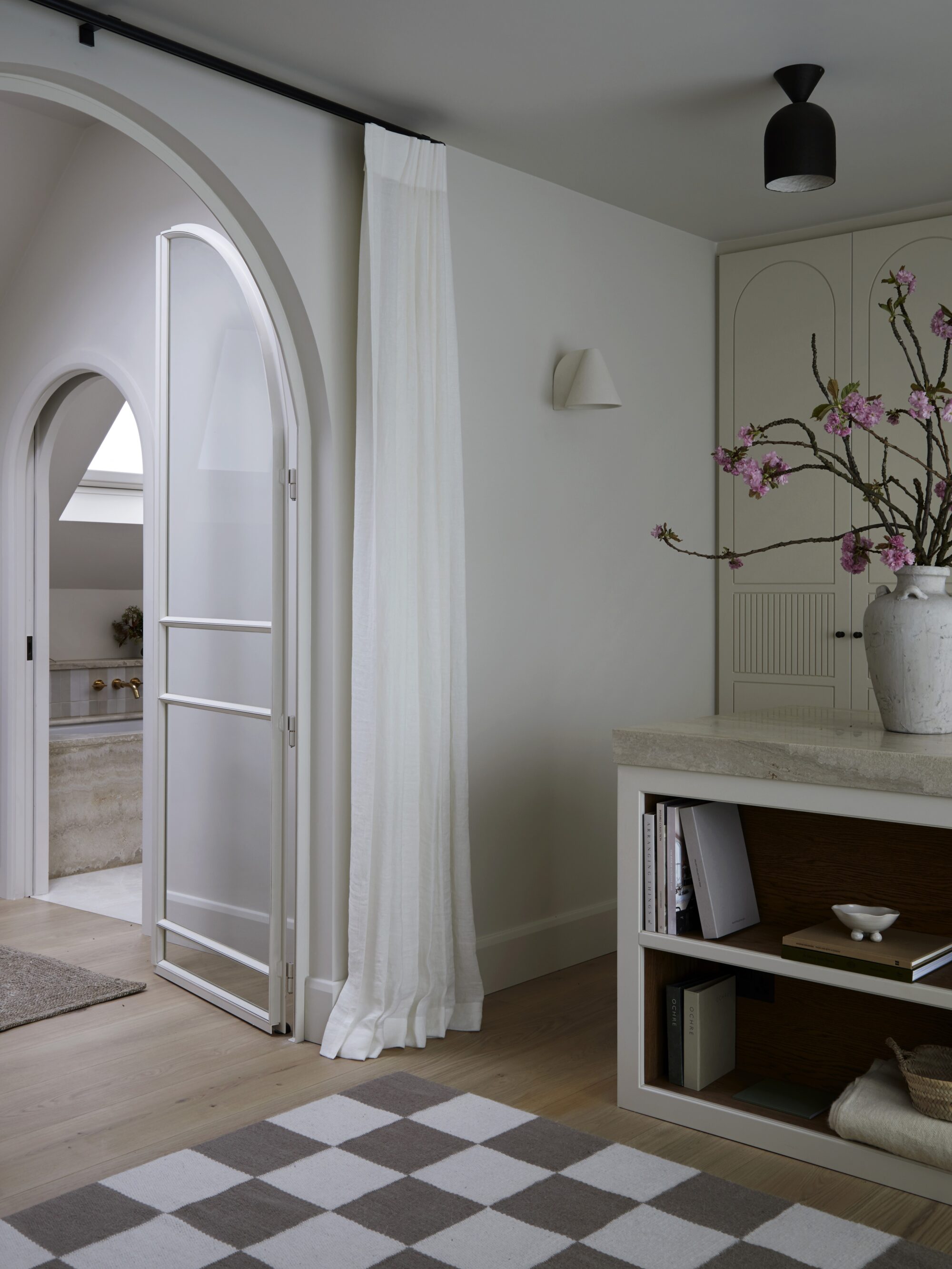 View through arched doorway from a bedroom looking into a landing and glimps of a bathroom to the right. The bedroom features white oak flooring with a monochrome scheme and checkered rug.