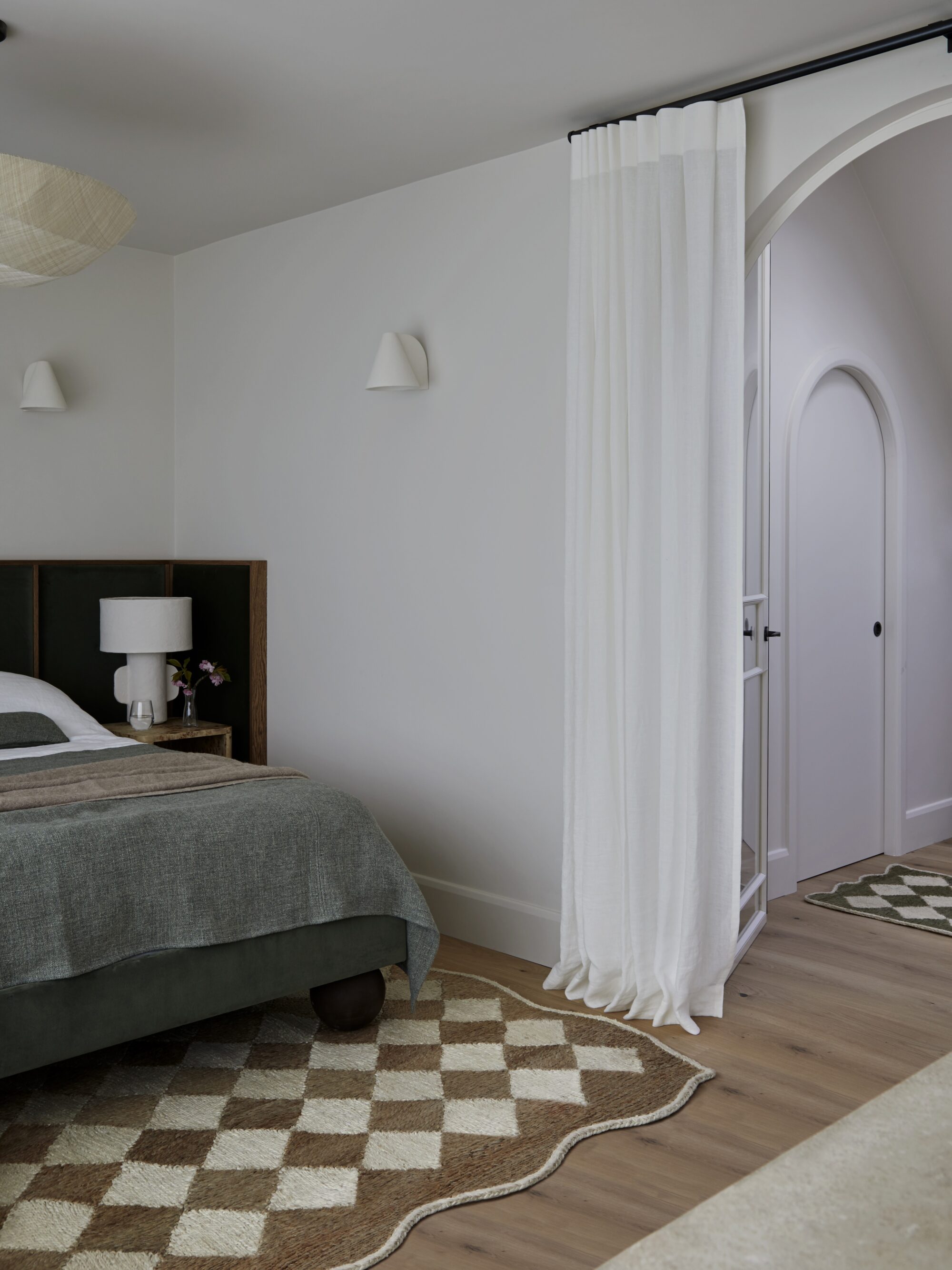 View through arched doorway from a bedroom with monochrome scheme, white oak flooring, a checkered rug and dark headboard. An arched doorway framed with white curtain offers a glimps into an adjacent landing. glimps of a bathroom to the right. The bedroom features white oak flooring with a monochrome scheme and checkered rug.