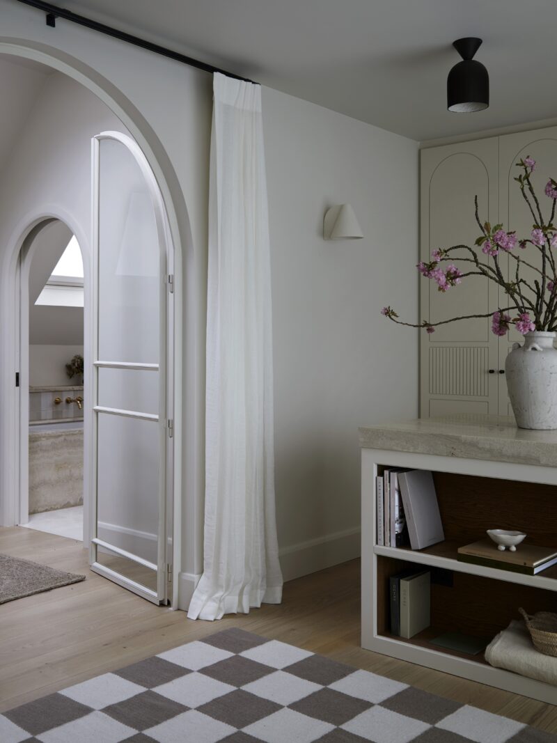 View through arched doorway from a bedroom looking into a landing and glimps of a bathroom to the right. The bedroom features white oak flooring with a monochrome scheme and checkered rug.